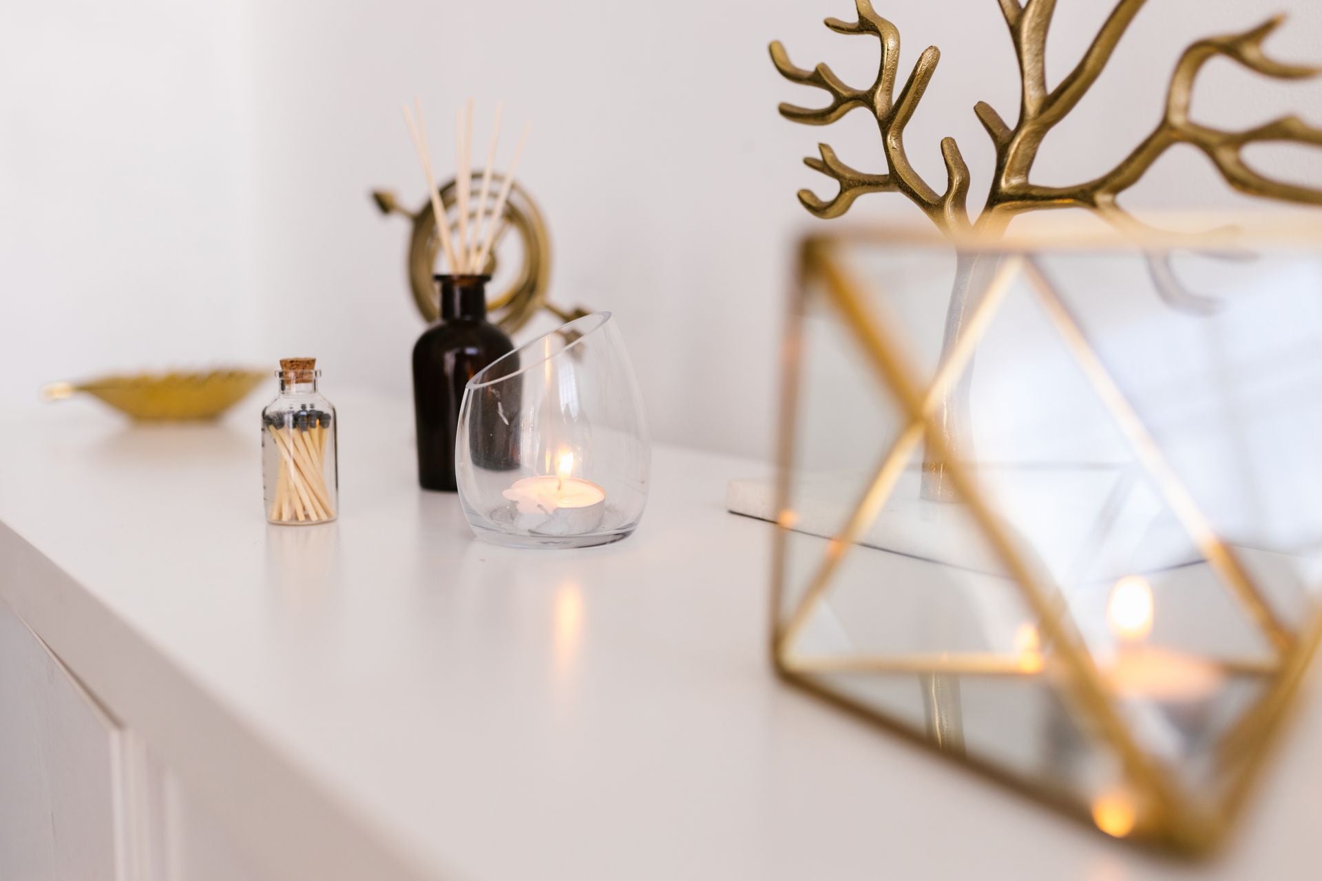 A styled interior tabletop scene with a white surface against a white wall, featuring a gold geometric candle holder in the foreground, a clear glass candle holder, a small bottle of matches, a dark bottle with reeds, and a gold decorative branch-like object.