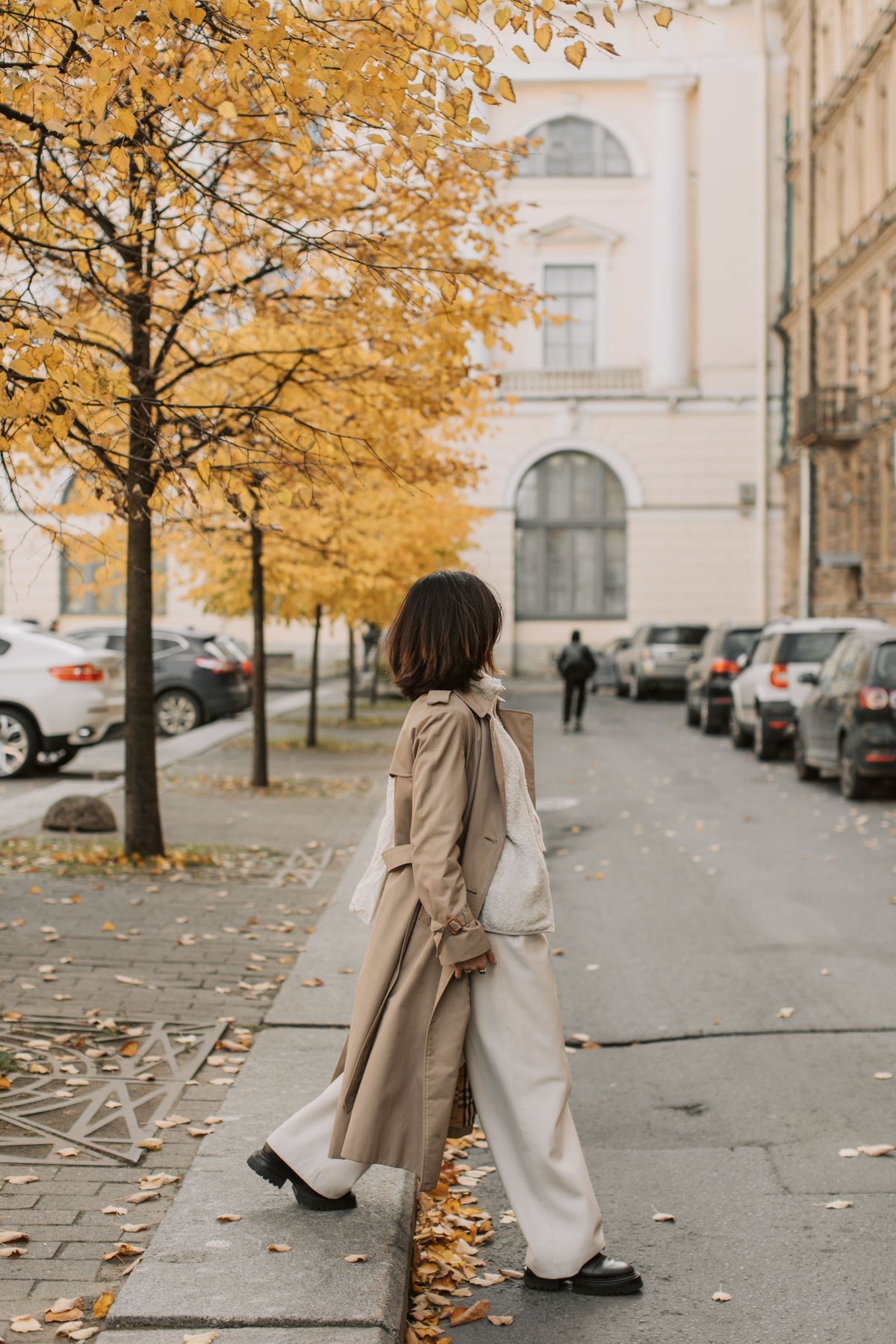 A woman wearing a beige trench coat over a white wide-leg dress walks along a city street lined with yellow autumn trees and parked cars.